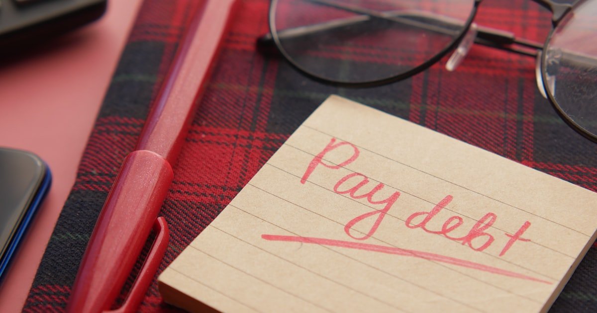 Handwritten note reading Pay debt next to a pen and glasses